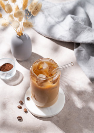 Ice latte coffee in a glass with milk on a light background with coffee beans, dry bouquet flowers and morning shadows. Trendy summer craft refreshing drink concept. Top view, copy space.の写真素材