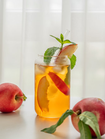 Ice tea with peach, mint in a glass on a white table near window with fresh fruits and sunlight close up. Refreshing summer drink concept for bar or cafe menu. Front view, copy space.の写真素材