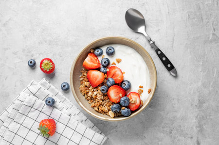 Natural yogurt with granola, blueberry and strawberry in a bowl on a light background with fresh berries, napkin and spoon. The concept of a healthy, nutritious and dietary breakfast. Top view, copy space.の写真素材
