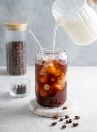 Milk pouring from a milk jug into a glass of iced coffee on a marble board on a light background with coffee beans. Trendy summer craft refreshing drink concept. Front view.の写真素材