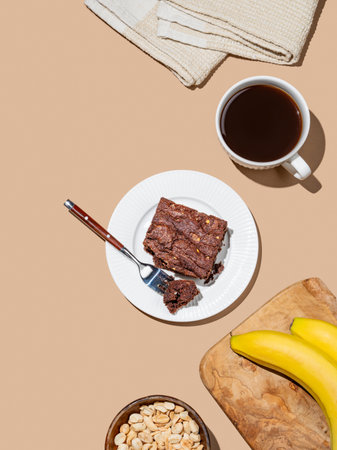 Chocolate banana brownie on a plate on a beige background with a cup of coffee, nuts and fresh fruits. Concept of dietary gluten-free baking for a cafe menu or advertising banner. Top view, copy spaceの写真素材