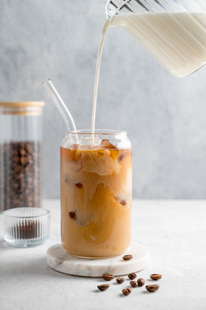 Milk pouring from a milk jug into a glass of iced coffee on a marble board on a light background with coffee beans. Trendy summer craft refreshing drink concept. Front view.の写真素材