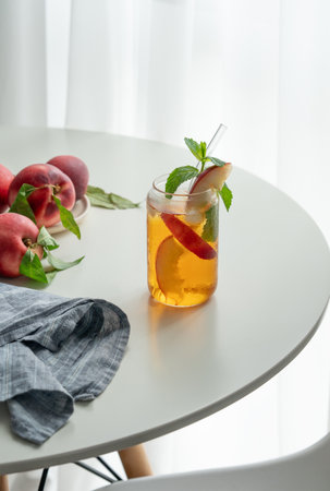 Ice tea with peach, mint in a glass on a white table near window with fresh fruits, napkin and sunlight. Refreshing summer drink concept for bar or cafe menu. Copy space.の写真素材