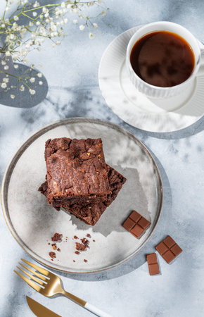 Chocolate brownie with nuts on a plate on a light background with cup of coffee, bouquet of flowers and morning shadows. Homemade sweet baking concept. Top view, copy space.の写真素材