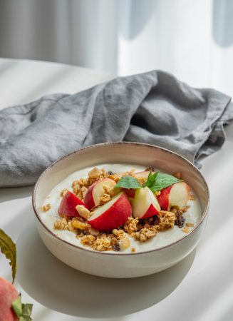 Natural greek yogurt with granola and peach in a bowl on a white table near window with napkin and morning shadows. Healthy and nutritious breakfast concept.の写真素材