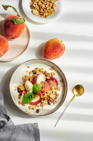 Natural greek yogurt with granola or muesli, peach and mint in a bowl on a white background with fresh fruits, spoon and morning shadows. Healthy and nutritious breakfast concept. Top view, copy spaceの写真素材