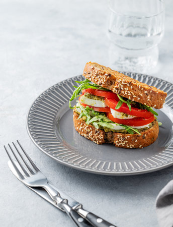 Sandwich of grain bread with fresh tomatoes mozzarella, arugula and pesto on a plate on a light background with glass of water and cutlery. Concept of healthy eating and dietary breakfast. Front view, copy space.の写真素材