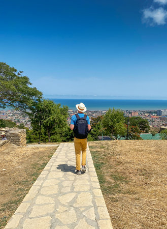 A man wearing a hat and carrying a backpack walks along a stone path toward a panoramic view of the city and the sea, with dry grass and trees under a bright blue sky. Dagestan, Derbentの写真素材