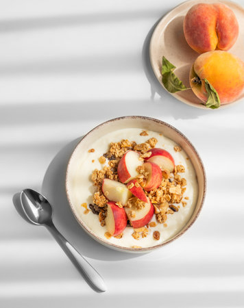 Natural greek yogurt with granola or muesli and peach in a bowl on a white background with spoon,...