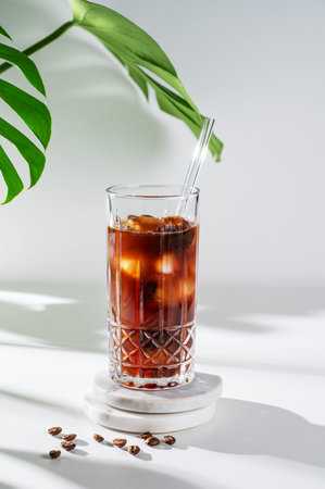 Cold brew coffee in a glass with ice on a light background with coffee beans, monstera flower leaves...
