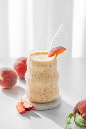 Smoothie with peach and banana in a glass on a white table near window with tropical fruit slices and morning sunlight. Healthy eating and detox diet concept. Front view, copy space.の写真素材