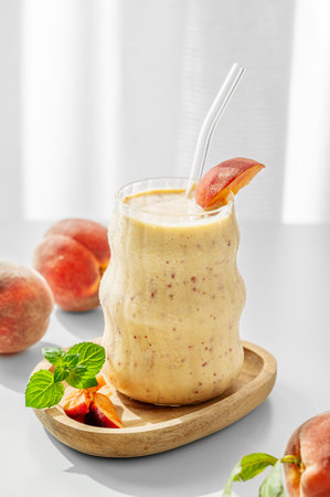 Smoothie with peach and banana in a glass on a wooden board on a white table near window with tropical fruit and morning sunlight. Healthy eating and detox diet concept. Front view, copy space.の写真素材