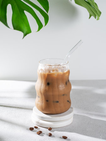 Ice latte coffee in a glass with milk and straw on a light background with coffee beans, monstera leaves and morning shadows. Summer refreshing trendy drink concept for advertising menu cafe. Front view, copy space.の写真素材