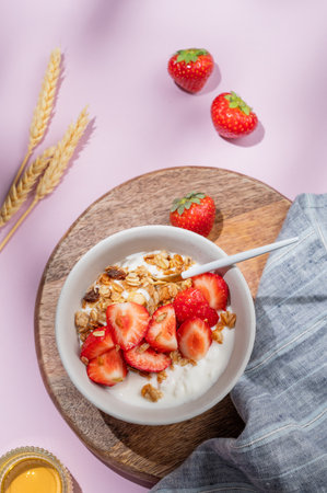 Granola or muesli with natural greek yogurt, strawberry and honey in a bowl on a wooden board on a violet background with wheat and shadows. Healthy and nutritious breakfast concept for advertising menu cafe or banner. Top view.の写真素材