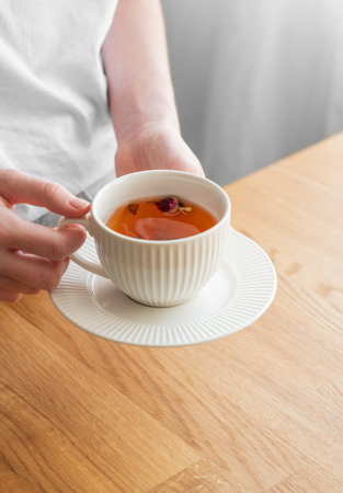 Woman holding white cup of tea with lemon and herbs on wooden tabletop near window with morning sunlight. Detox breakfast and healthy eating concept. Copy space.の写真素材