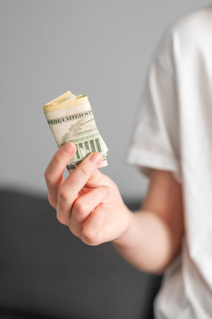 Female hand holding a twisted stack of US dollar bills on a gray background indoors. Finance,...