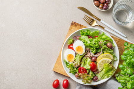 Tuna salad with olives, lettuce, tomatoes, eggs and lemon in a plate on a wooden board on a light background with glass of water and cutlery. The concept of healthy eating and the Mediterranean diet. Top view, copy space.の写真素材