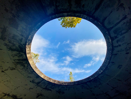 A round hole in a cracked concrete ceiling reveals a view of blue sky with clouds and treetops, taken from below in natural daylight. Tskaltubo, Georgia.の写真素材