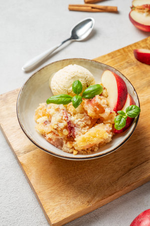 Peach crumble or cobbler with vanilla ice cream scoops and cinnamon in a plate on a wooden board on a light background with fresh fruits and spoon. Summer homemade refreshing dessert concept.の写真素材