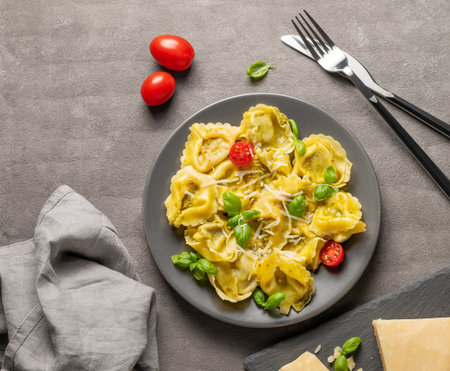 Tortellini pasta with parmesan, pesto and basil in a plate on a dark background with cheese, cutlery and tomato. Traditional Italian homemade food concept in rustic style. Top view, copy space.の写真素材