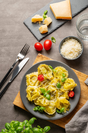 Tortellini pasta with parmesan, pesto and basil in a plate on a wooden board on a dark background with cheese, herb, tomato and cutlery. Traditional Italian homemade food concept in rustic style. Top view, copy space.の写真素材