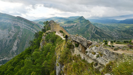 The ruins of a stone fortress sit atop a rocky mountain ridge with green valleys down below in...