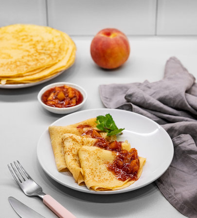 Pancakes with peach jam on a plate on a light kitchen brick background with fresh fruits, napkin and...