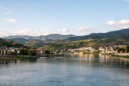 A small coastal town with houses and buildings, located by a calm river, surrounded by wooded hills...