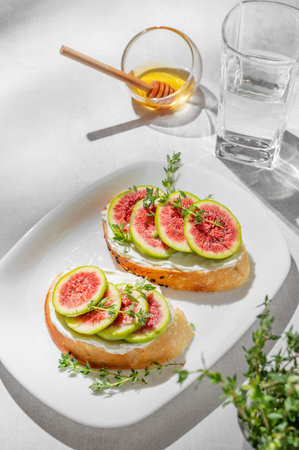 Sweet toasts with fresh figs, cream cheese, thyme, and honey on a light background. Concept of a delicious dietary appetizer of bruschetta and fruit on a plate with glass of water and shadows. Top view, copy space.の写真素材