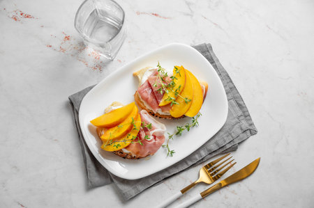 Toast with peach, cream cheese, ham and honey on a plate on a marble background with thyme and glass of water. Healthy diet snack concept for a advertising menu or cafe banner. Top view, copy space.の写真素材
