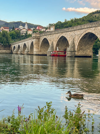 An ancient stone bridge with arches spanning the tranquil Drina River with wild flowers in the foreground and lush green hills and houses in the background. Heritage and developing tourism concept. Visegrad, Bosnia and Herzegovina.の写真素材