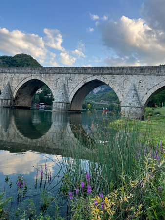 An ancient stone bridge with arches spanning the tranquil Drina River with wild flowers in the foreground and lush green hills and houses in the background. Heritage and developing tourism concept. Visegrad, Bosnia and Herzegovina.の写真素材