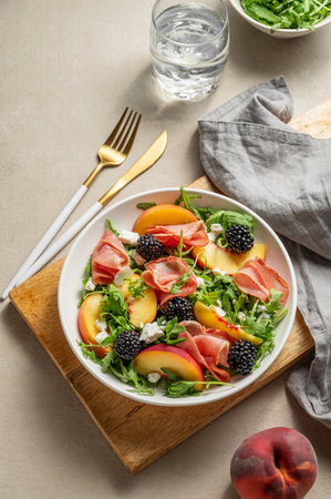 Fresh salad with peaches, arugula, feta, prosciutto and berries in a plate on a wooden board on a light background with glass of water and napkin. Summer dietary healthy food concept. Top view.の写真素材