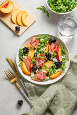 Fresh salad with peaches, arugula, feta, prosciutto and berries in a plate on a light background with glass of water and napkin. Summer dietary healthy snack concept for advertising a menu. Top view.の写真素材
