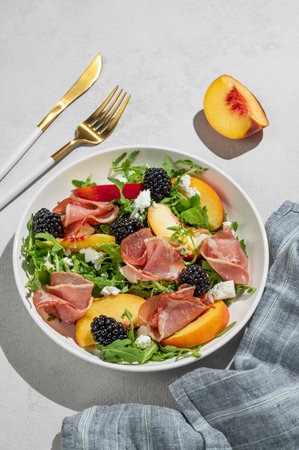 Fresh salad with peaches, arugula, feta, prosciutto and berries in a white plate on a light background with shadows. Summer dietary healthy snack concept for advertising a menu. Top view, copy space.の写真素材