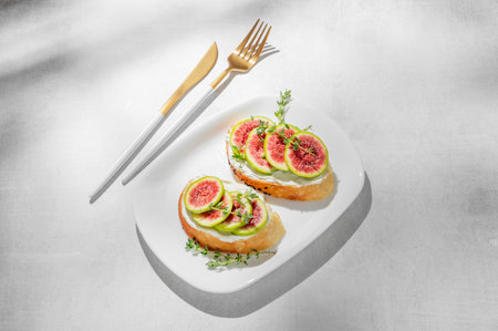 Sweet toasts with fresh figs, cream cheese, thyme, and honey on a light background. Concept of a delicious dietary appetizer of bruschetta and fruit on a plate with cutlery and shadows. Top view, copy space.の写真素材