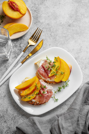Bruschetta with peach, cream cheese, prosciutto and honey on a plate on a gray background with thyme, napkin and cutlery. Healthy diet snack concept for a advertising menu or cafe banner. Top view, copy space.の写真素材