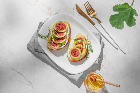 Sweet toasts with fresh figs, cream cheese, thyme, and honey on a marble background. Concept of a delicious dietary appetizer of bruschetta and fruit on a plate with cutlery and shadows. Top view, copy space.の写真素材