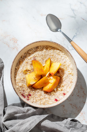 Oatmeal with peaches and nuts  in a bowl on a marble background with honey, shadows, spoon and napkin. Concept for a healthy vegan dish or breakfast snack. Top view, copy space.の写真素材