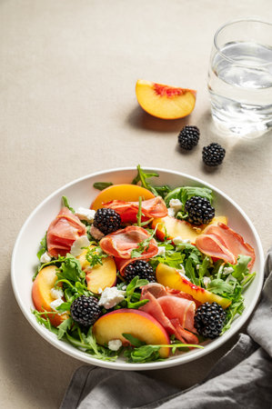 Fresh salad with peaches, arugula, feta, prosciutto and berries in a plate on a light background with glass of water and napkin. Summer dietary healthy food concept. Copy space.の写真素材