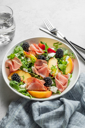Fresh salad with peaches, arugula, feta, prosciutto and berries in a plate on a light background with glass of water. Summer dietary healthy snack concept for advertising a menu. Copy space.の写真素材