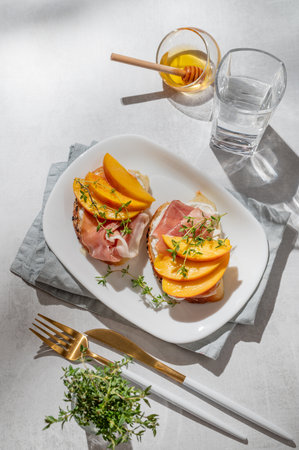 Toast with peach, cream cheese, ham and honey on a plate on a light background with thyme, glass of water and shadows. Healthy diet snack concept for a menu or cafe banner. Top view, copy space.の写真素材