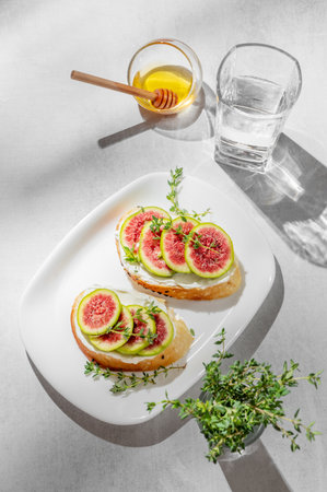 Sweet toasts with fresh figs, cream cheese, thyme, and honey on a light background. Concept of a delicious dietary appetizer of bruschetta and fruit on a plate with glass of water and shadows. Top view, copy space.の写真素材