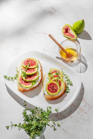 Sweet toasts with fresh figs, cream cheese, thyme, and honey on a marble background. Concept of a delicious dietary appetizer of bruschetta and fruit on a plate with shadows. Top view, copy space.の写真素材
