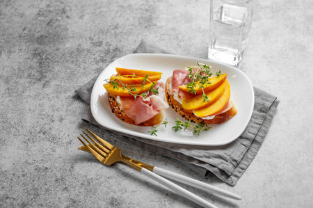 Sandwich with peach, cream cheese, prosciutto and honey on a plate on a gray background with thyme...