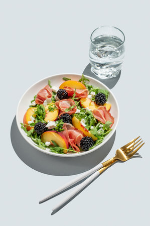 Salad with peaches, arugula, feta, prosciutto and berries in a plate on a blue background with glass of water, cutlery and shadow. Summer healthy food concept for advertising menu cafe. Copy space.の写真素材