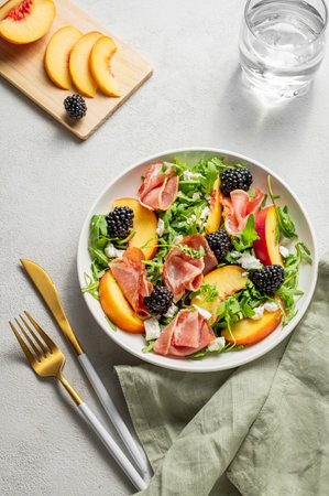 Fresh salad with peaches, arugula, feta, prosciutto and berries in a plate on a light background with glass of water and napkin. Summer dietary healthy snack concept for advertising a menu. Top view.の写真素材
