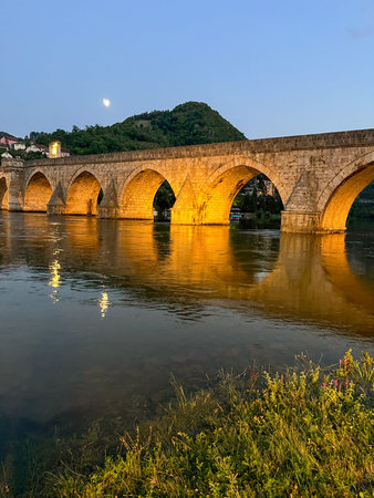 Historic stone bridge with illuminated arches over the Drina River at dusk. Grassy bank, moon and wooded hill in the background. Visegrad, Bosnia and Herzegovina.の写真素材