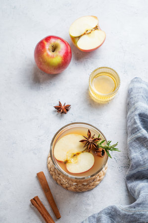 Hot apple cider with cinnamon, anise, and rosemary in glass on a light background with honey, fresh fruits and napkin. Concept a traditional, spicy, warming drink for fall and winter. Top view, copy space.の写真素材