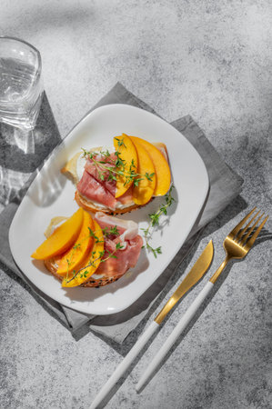 Bruschetta with peach, cream cheese, prosciutto and honey on a plate on a gray background with thyme, cutlery and shadows. Healthy diet snack concept for a advertising menu or cafe banner. Top view, copy space.の写真素材